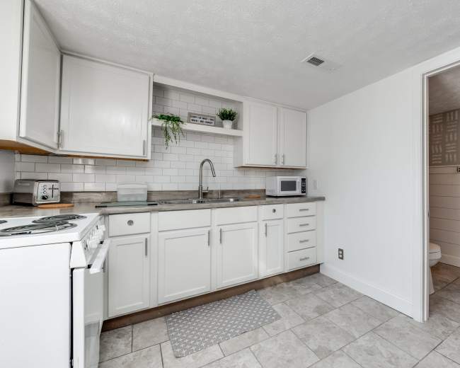 A clean kitchen with white cabinetry, a sink, a microwave, and tiled flooring, adjacent to a bathroom area.