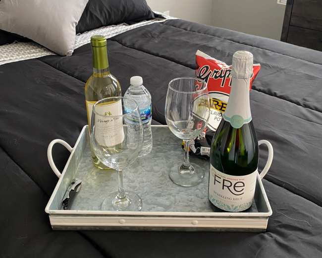 A tray holds a bottle of sparkling wine, a bottle of white wine, two wine glasses, a bottle of water, and a bag of chips on a made bed with dark bedding.