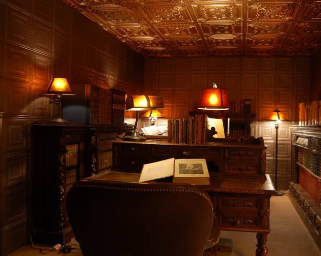 A traditional library features a dark wood desk, ornate bookshelves filled with books, and soft lighting from lamps on both the desk and shelves.