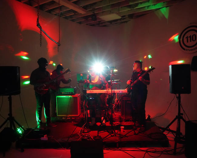 A band performs on stage with colorful lights illuminating the space, surrounded by amplifiers and musical instruments.