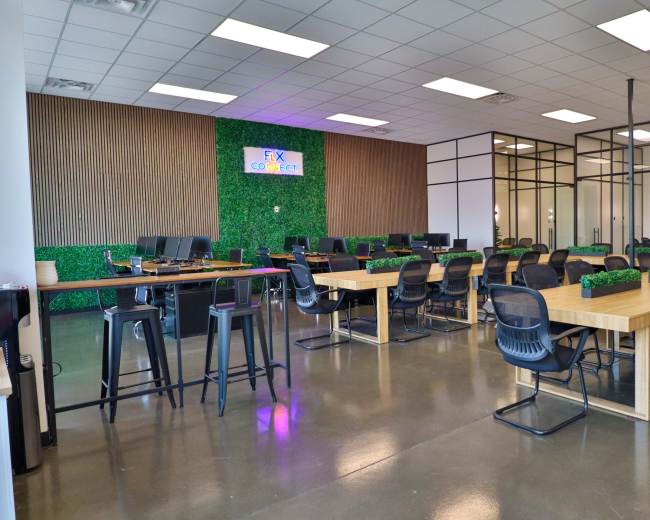 The image shows a spacious coworking area with large tables, black office chairs, and a green wall in the background featuring a sign that reads "FX Coworking."