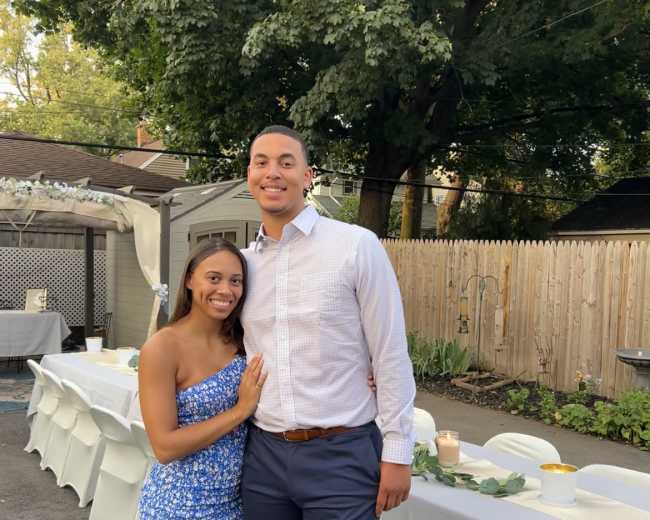 A couple stands together in a garden setting with a dining area in the background, surrounded by trees and chairs.