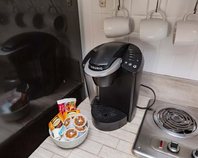 A Keurig coffee maker sits on a countertop next to a bowl of coffee pods and a stovetop with hanging mugs in the background.