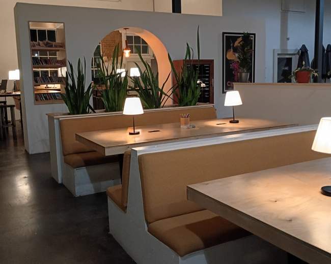 The image shows a modern cafe interior with wooden tables, cushioned benches, and potted plants, illuminated by soft table lamps.