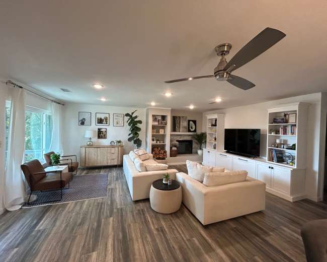The image shows a modern living room with a seating area that includes two white sofas and a black table, alongside a window with light curtains and a television mounted on a white cabinet.