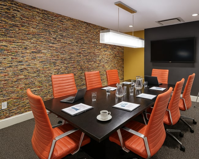 A modern conference room features a large table surrounded by eight orange chairs, a wall adorned with textured material, and a television displaying a presentation in the background.