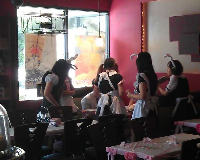 A group of waitresses in maid outfits stands around a table in a cafe, interacting with customers.