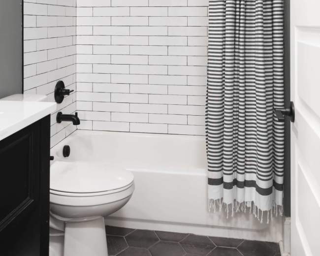 A bathroom features a white tiled shower area with a black and white striped curtain, a modern toilet, and a dark vanity with a hexagon tile floor.