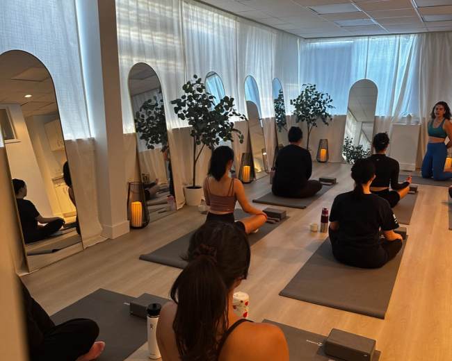 A group of individuals is seated on yoga mats in a well-lit studio with mirrors and plants, preparing for a class.