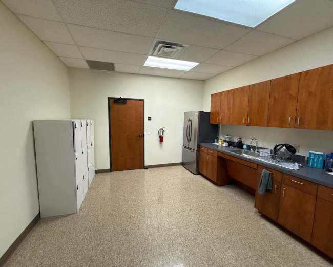 The image shows a break room with wooden cabinets, a refrigerator, and a row of lockers along one wall.