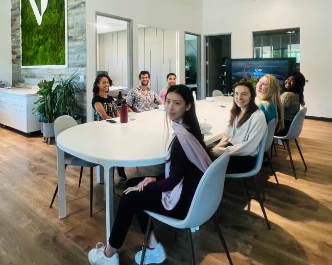 A group of people is sitting around a large, round table in a well-lit modern office with greenery on the walls and various plants around the room.
