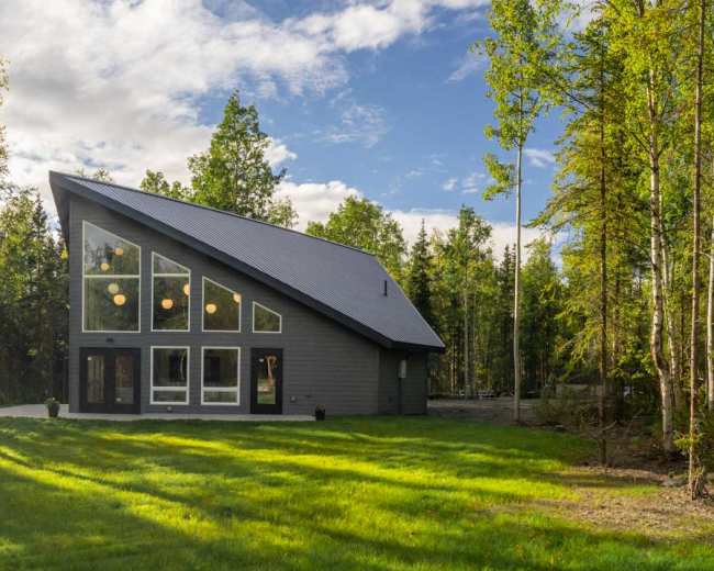 A modern, A-frame house with large windows is situated in a forest clearing, surrounded by greenery and sunlight filtering through the trees.