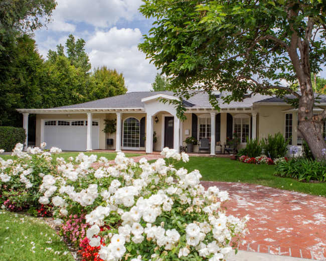 A single-story house with a front porch, surrounded by well-maintained gardens and a brick pathway.