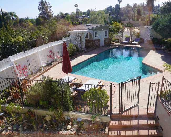 The image shows a backyard pool area with a fenced-in pool, a lounging area, and surrounding greenery.