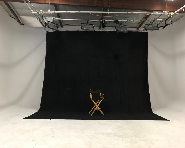 The image shows a photography studio with a black backdrop and a single wooden director's chair positioned in the center.