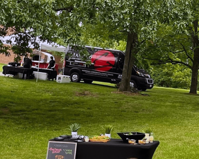 A food truck is parked next to a canopy with tables set up on a grassy lawn.