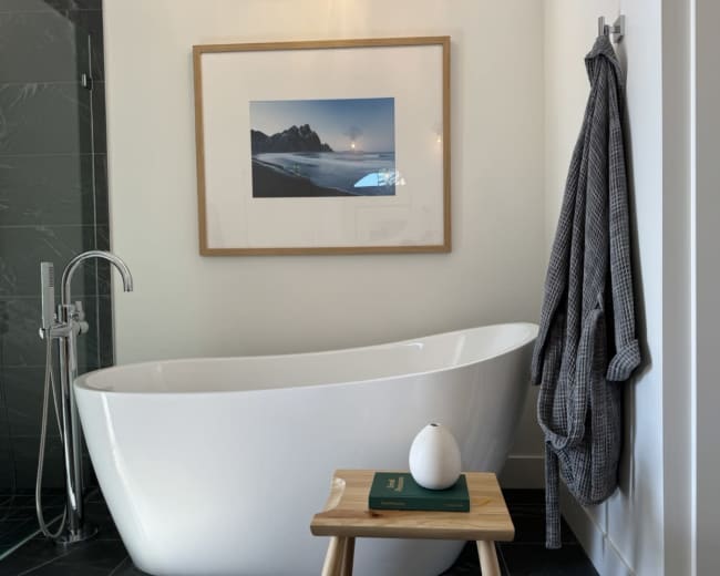 A freestanding white bathtub is positioned next to a framed landscape photograph, accompanied by a wooden stool holding a decorative item and a book.