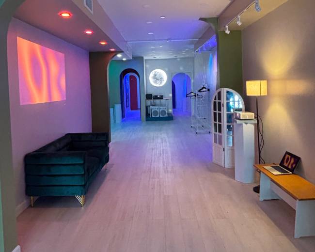 The image shows a modern interior space with arched doorways, colorful lighting, and a long hallway leading to a circular mirror at the end.