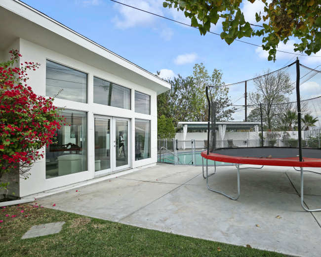 The image shows a modern house with large windows adjacent to a trampoline and a swimming pool surrounded by concrete and greenery.