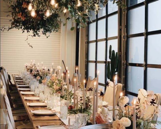 A long dining table, set with candles and floral arrangements, is decorated with hanging plants and pendant lights in a modern venue.