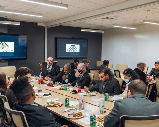 A group of people in business attire are seated around a long table, eating and engaged in conversation during a meal in a conference room.