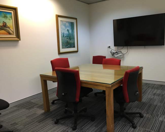 A conference room features a glass-top table surrounded by four red chairs, with two framed paintings on the walls and a mounted television.