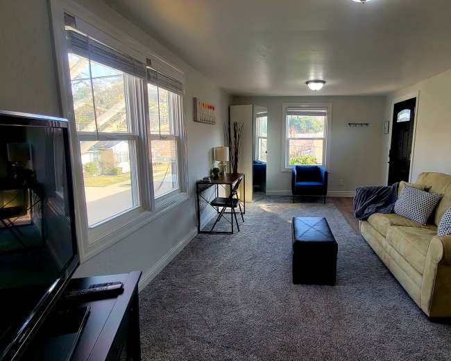 The room features a beige sofa, a dark ottoman, a television, and large windows providing natural light, with a mirror and a chair visible in the background.