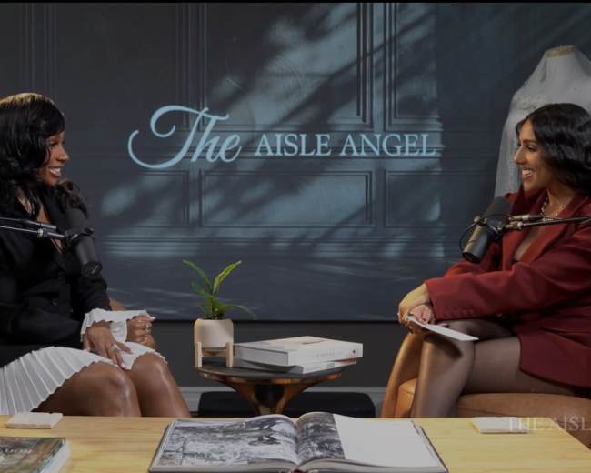 Two women sit across from each other at a table adorned with books and a plant, engaged in conversation in a well-lit studio setting with a backdrop featuring the text "The Aisle Angel."