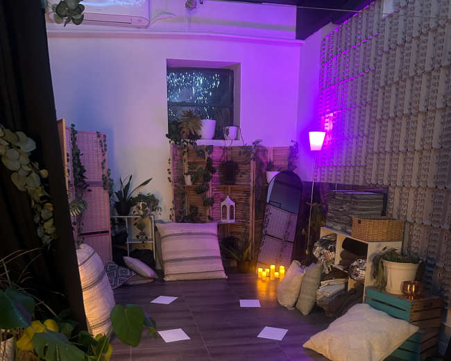 The image shows a cozy indoor space with various cushions, plants, and candles arranged around a central floor area, illuminated by purple lighting.