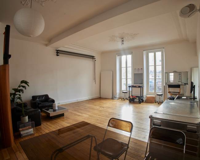 The image shows a spacious, minimalist room with hardwood flooring, a black leather sofa, a table with transparent surface, and large windows letting in natural light.