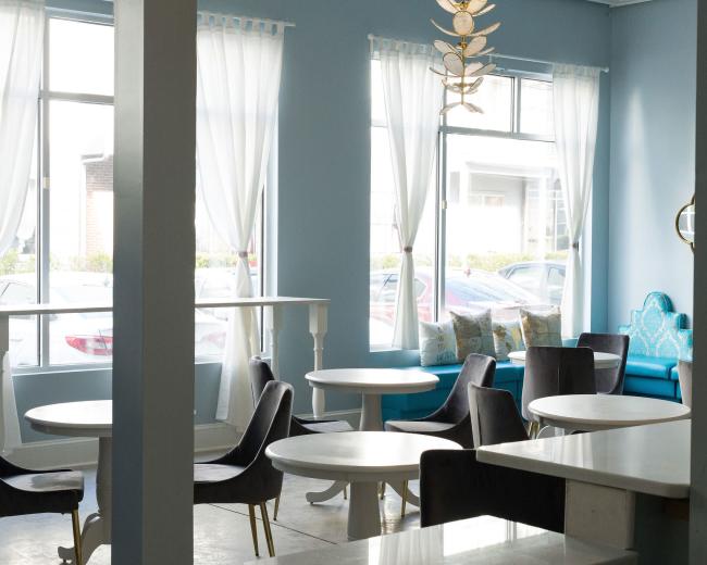 A brightly lit café interior with blue walls, large windows, and a mix of round tables and upholstered chairs.