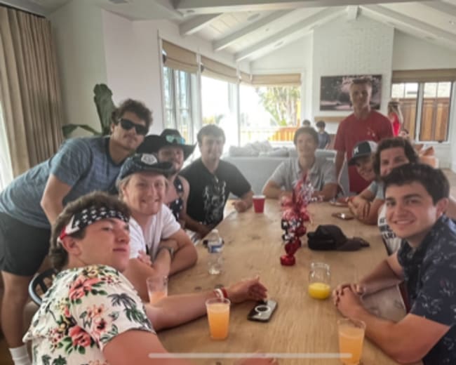 A group of people is gathered around a wooden table in a bright room, enjoying drinks and engaging with each other.