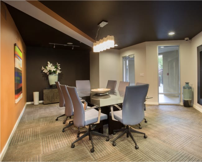 The image shows a modern conference room with a glass-top table surrounded by six chairs, a decorative vase, and an abstract painting on the wall.