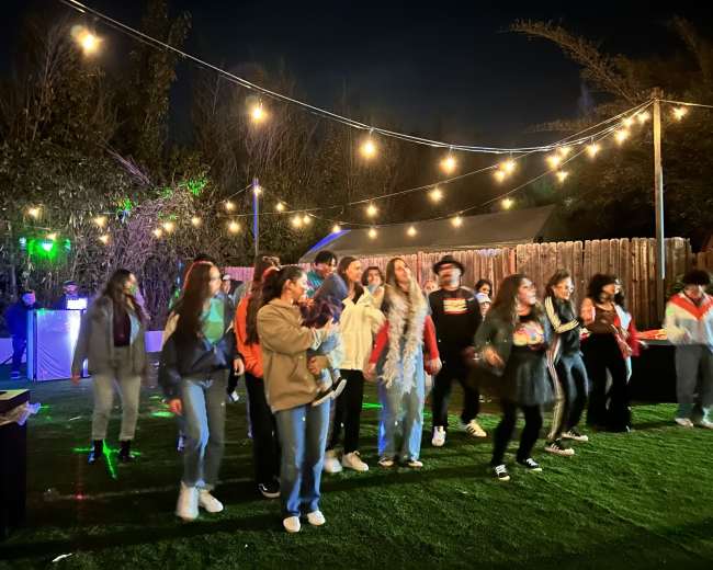 A group of people is dancing in an outdoor setting illuminated by string lights at night.