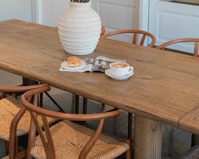 A large white vase with branches sits on a wooden table next to a cup of coffee and a pastry beside an open magazine.