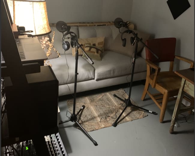 The image shows a recording studio setup featuring a sofa, two microphones on stands, a wooden chair, and a lamp next to a rug.