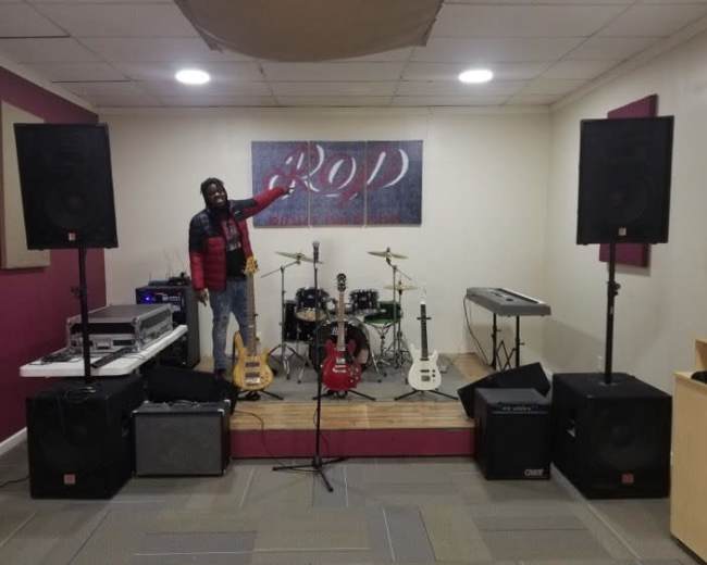 A man stands on a wooden stage in a music rehearsal space surrounded by musical instruments and audio equipment.