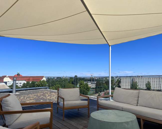 The image shows a rooftop terrace with beige outdoor furniture, a round green table, and a large umbrella, overlooking a cityscape under a clear blue sky.