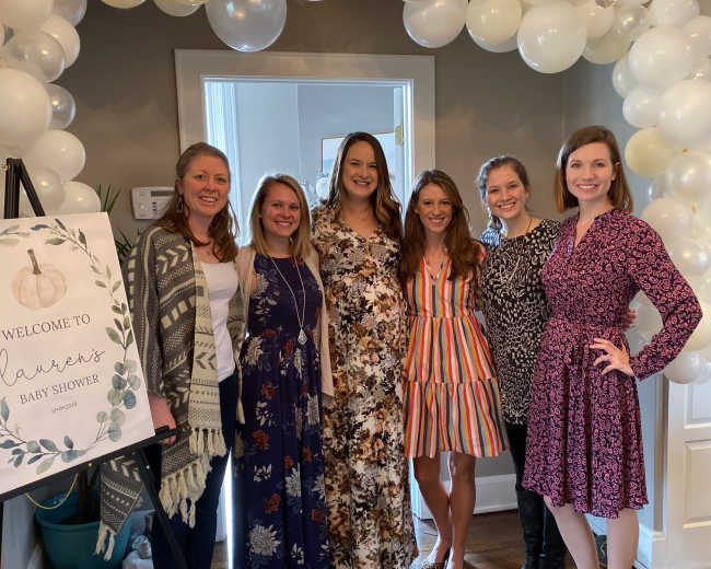 A group of six women stands together in a decorated room, posing in front of a balloon arch and a sign that reads "Welcome to Lauren's Baby Shower."