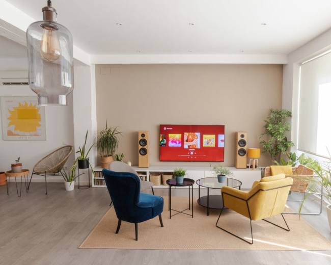 A modern living room features two chairs, a coffee table, and a large TV mounted on the wall, surrounded by plants and decorative items.