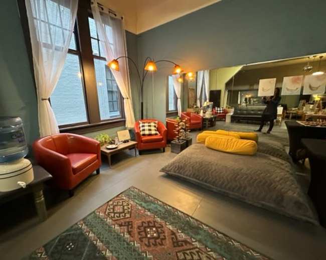 The image shows a cozy, well-lit lounge area with two red armchairs, a large mirror, and a water cooler, accompanied by a patterned rug and soft cushions on the floor.