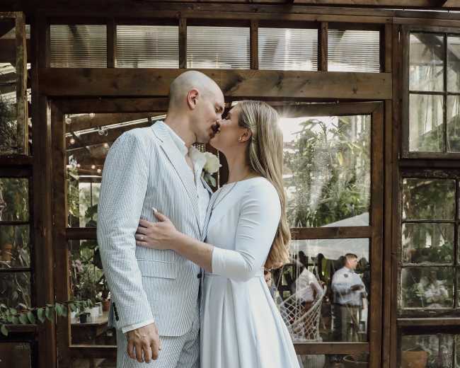 A couple shares a kiss in an indoor garden setting, surrounded by glass walls and greenery.