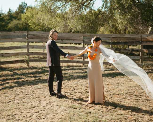 A couple stands in a grassy field near a wooden fence, with the woman wearing a form-fitting dress and a floral necklace, while her veil flows in the breeze.