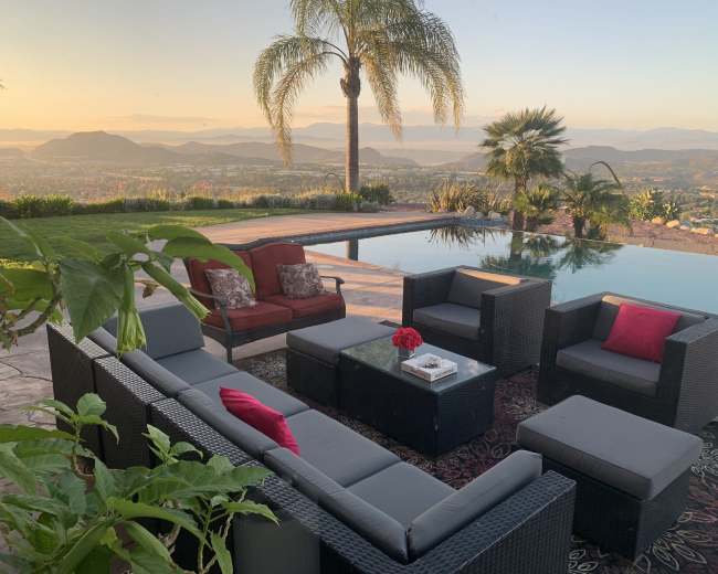A modern outdoor seating area with black wicker furniture and red accents overlooks a scenic landscape at sunset.