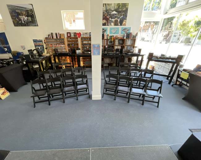 The image shows a room with rows of black chairs arranged in front of a small stage, surrounded by shelves filled with books and music equipment.