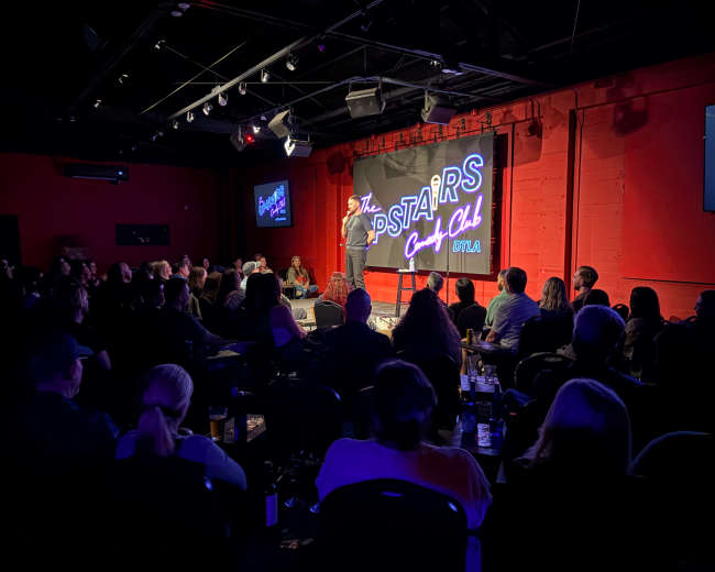A stand-up comedian performs on stage in a dimly lit comedy club while an audience watches attentively.
