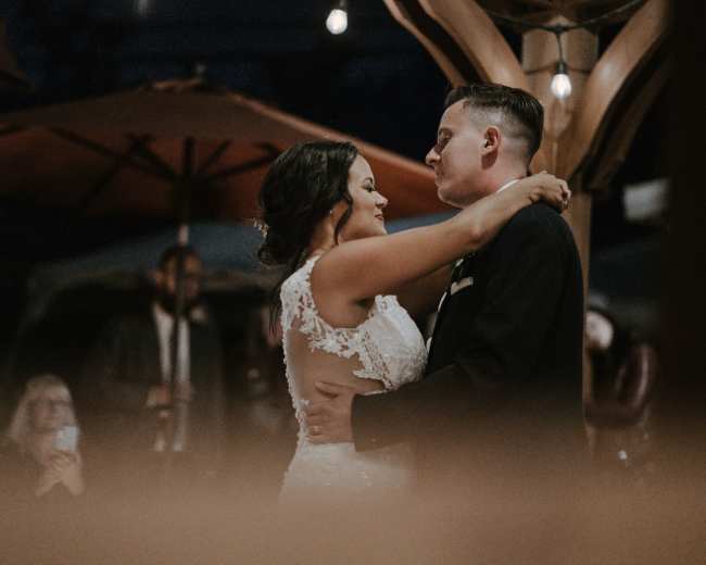 A couple dances closely under dim lights at an outdoor venue, surrounded by guests.