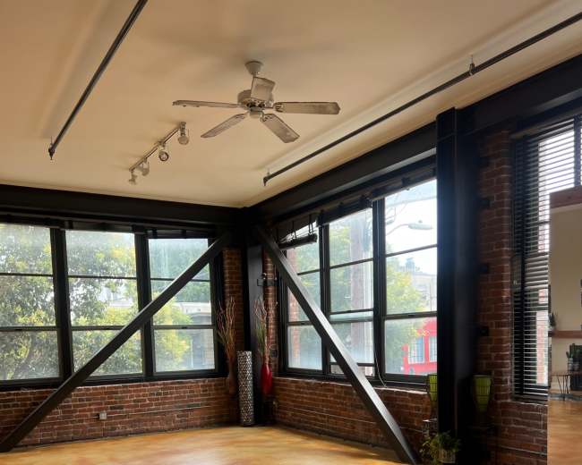 The image shows an empty room with large windows, exposed brick walls, and a concrete floor.
