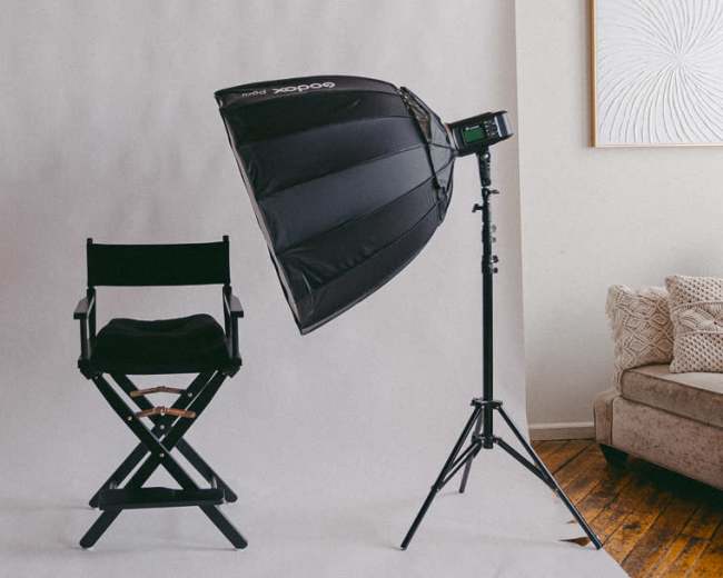The image shows a photography setup featuring a black director's chair beside a light modifier mounted on a stand, all against a white backdrop.