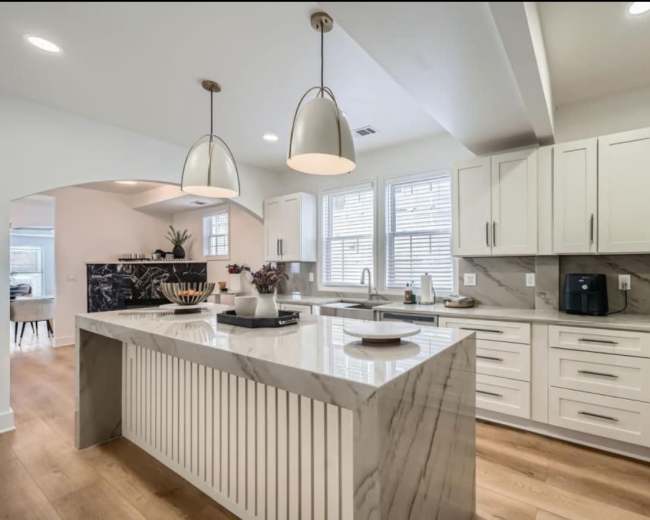 The image shows a modern kitchen with a large marble-topped island, white cabinetry, and bright natural light coming through the windows.